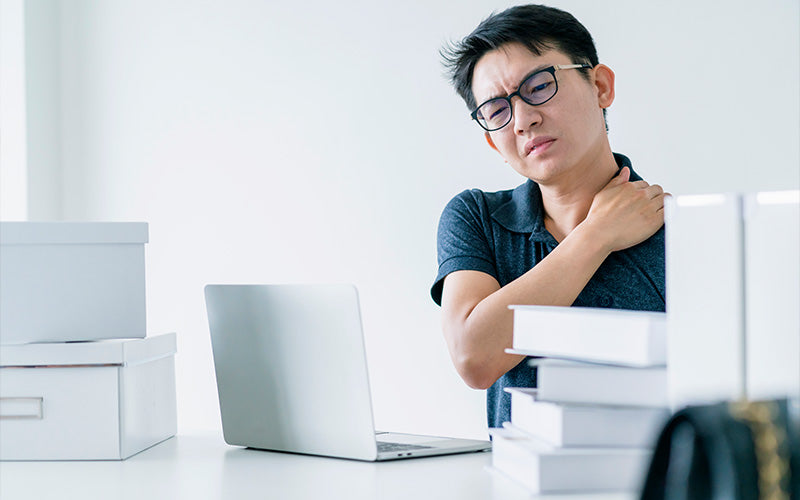 A man sitting at his desk, grimacing and holding his shoulder in pain