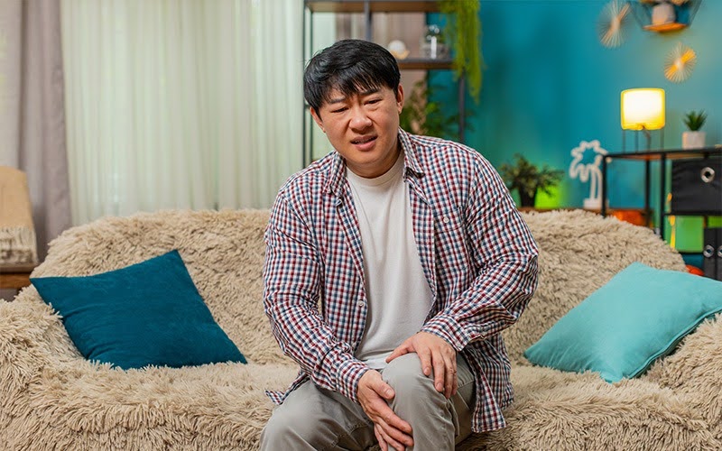 Man sitting on a couch expressing discomfort and holding his knee.